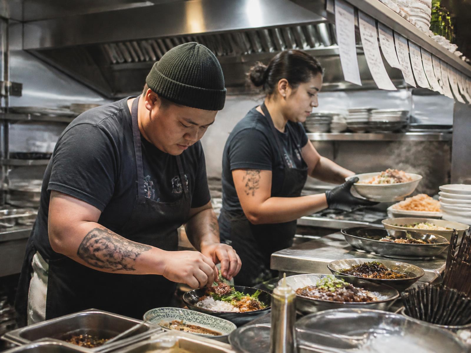 Two cooks assembling and plating dishes at a stainless steel kitchen pass during service, with order tickets on the rail above them