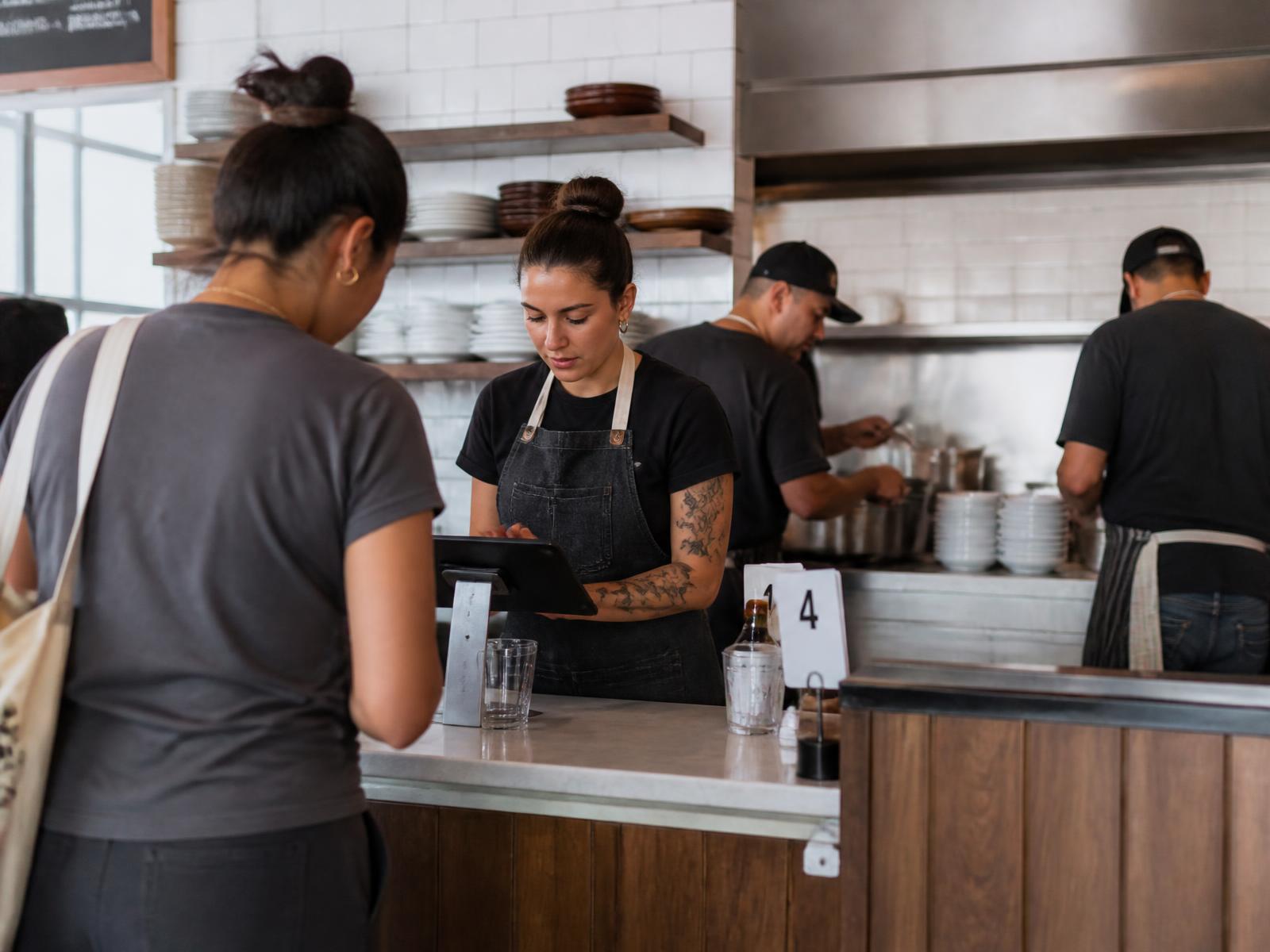 Restaurant staff member taking an order at the counter on a tablet point-of-sale terminal while cooks work at the open kitchen pass behind her