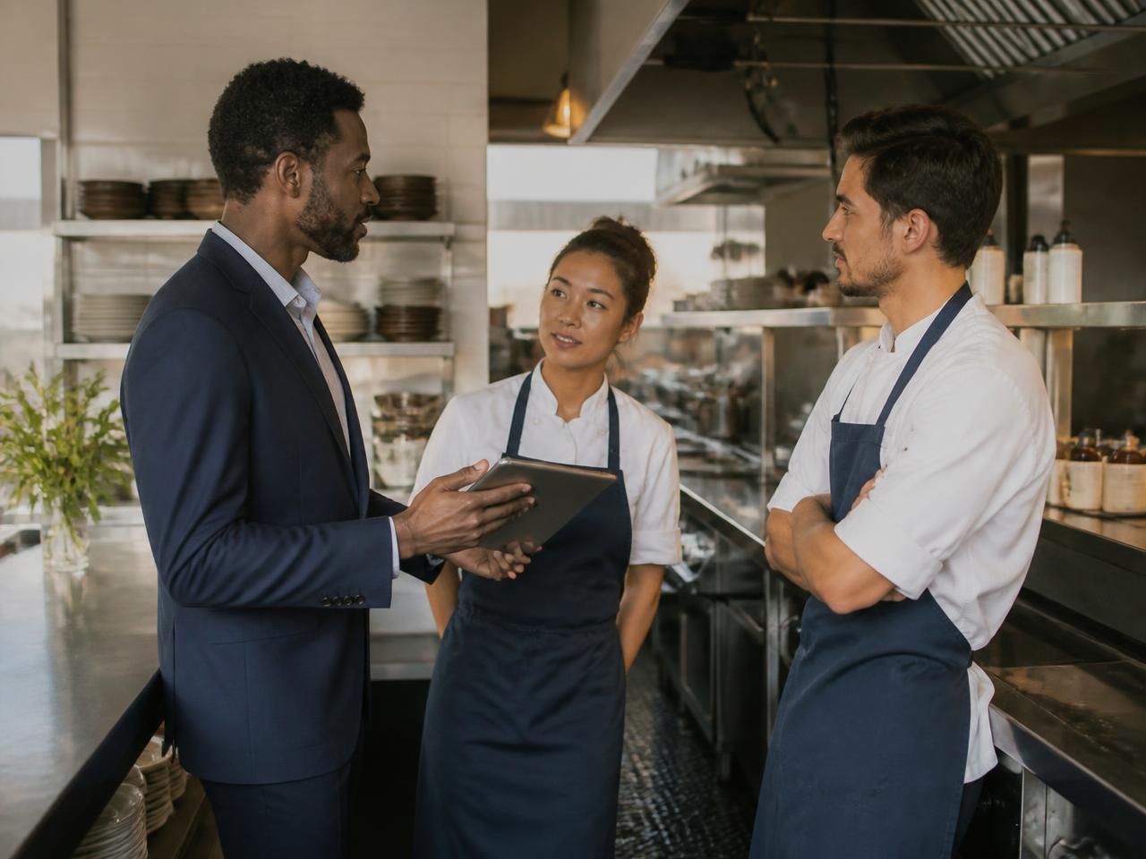 Restaurant manager with a tablet talking with two team members near the pass of a commercial kitchen